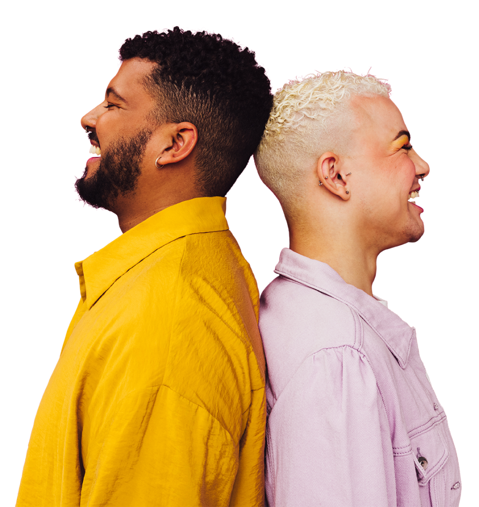 Two people stand back-to-back, smiling broadly. The person on the left has short dark curly hair and a beard, wearing a yellow shirt. The person on the right has short light curly hair and earrings, wearing a light purple jacket.