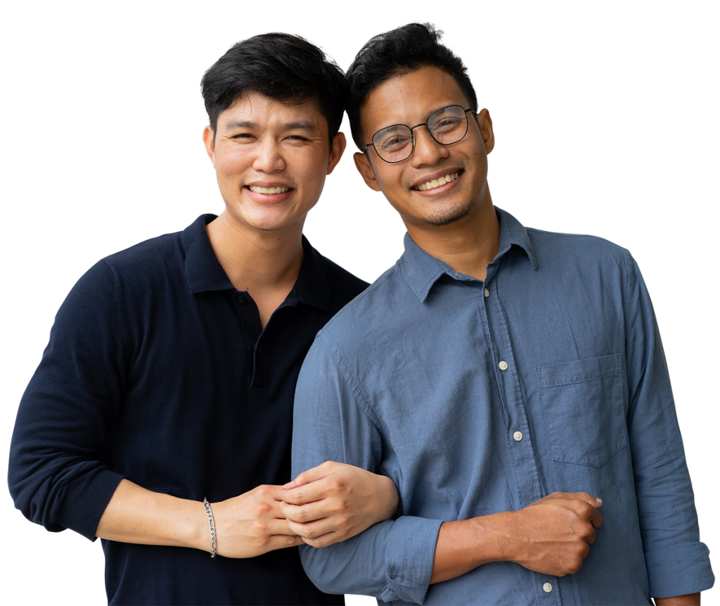 Two men stand close together, smiling warmly at the camera. The man on the left has short dark hair and wears a black long-sleeved shirt, while the man on the right has glasses, short curly hair, and wears a blue button-up shirt. They appear cheerful and friendly.
