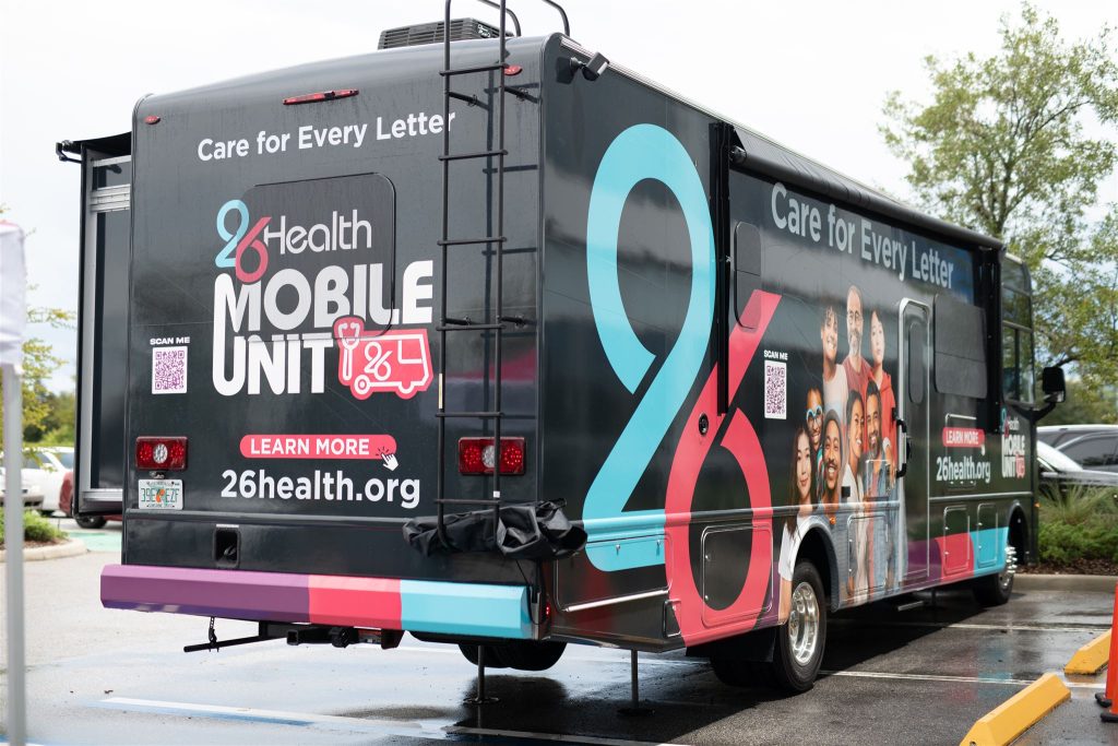 A mobile health unit van parked outdoors. The van features colorful branding with text reading "26Health Mobile Unit" and "Care for Every Letter." Images of diverse individuals and a QR code are visible on the side. The website "26Health.org" is also displayed.