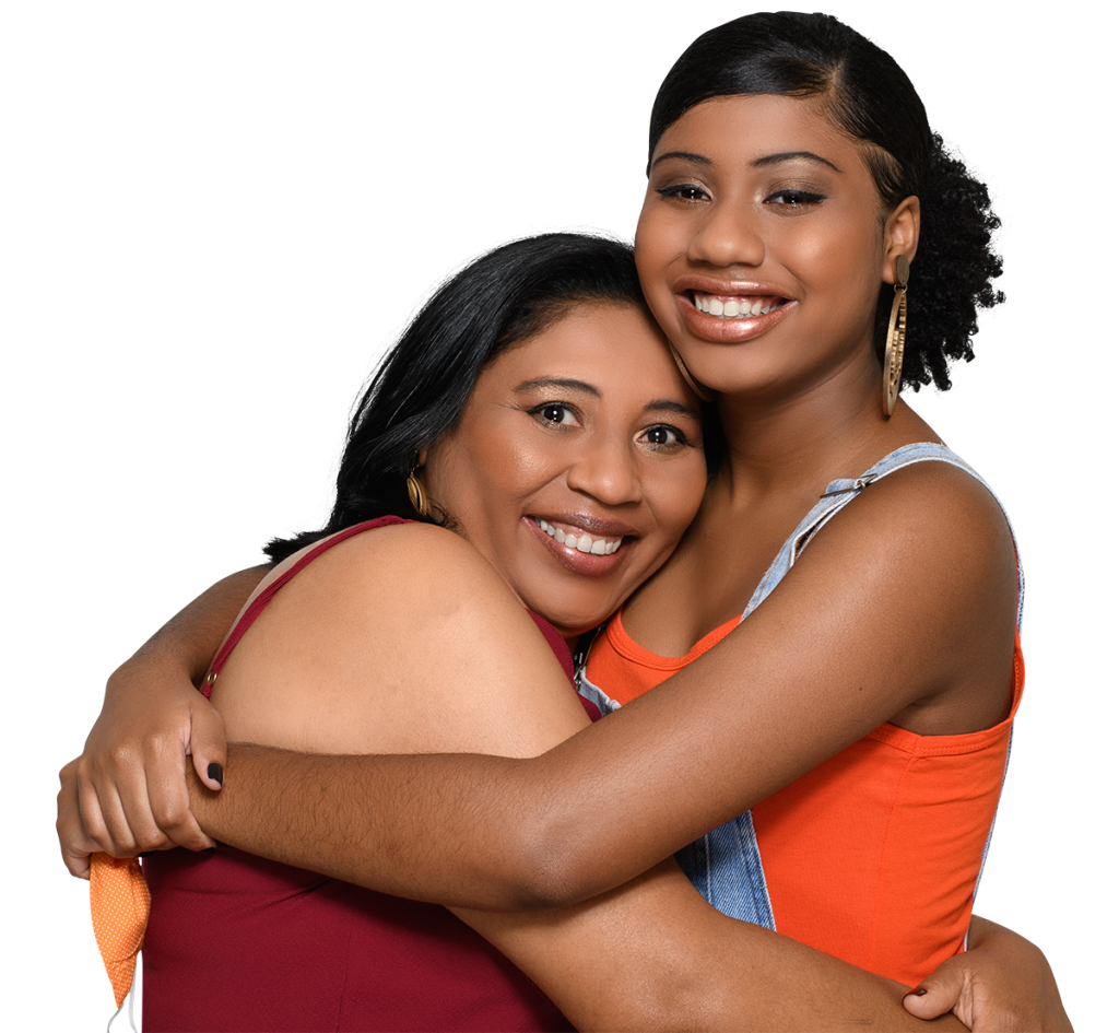 Family and Relationship Therapy. Two women are smiling and hugging each other. The woman on the left has shoulder-length black hair and wears a maroon top. The woman on the right has curly hair pulled back, gold hoop earrings, and wears an orange top with denim overalls. Both look happy and affectionate.
