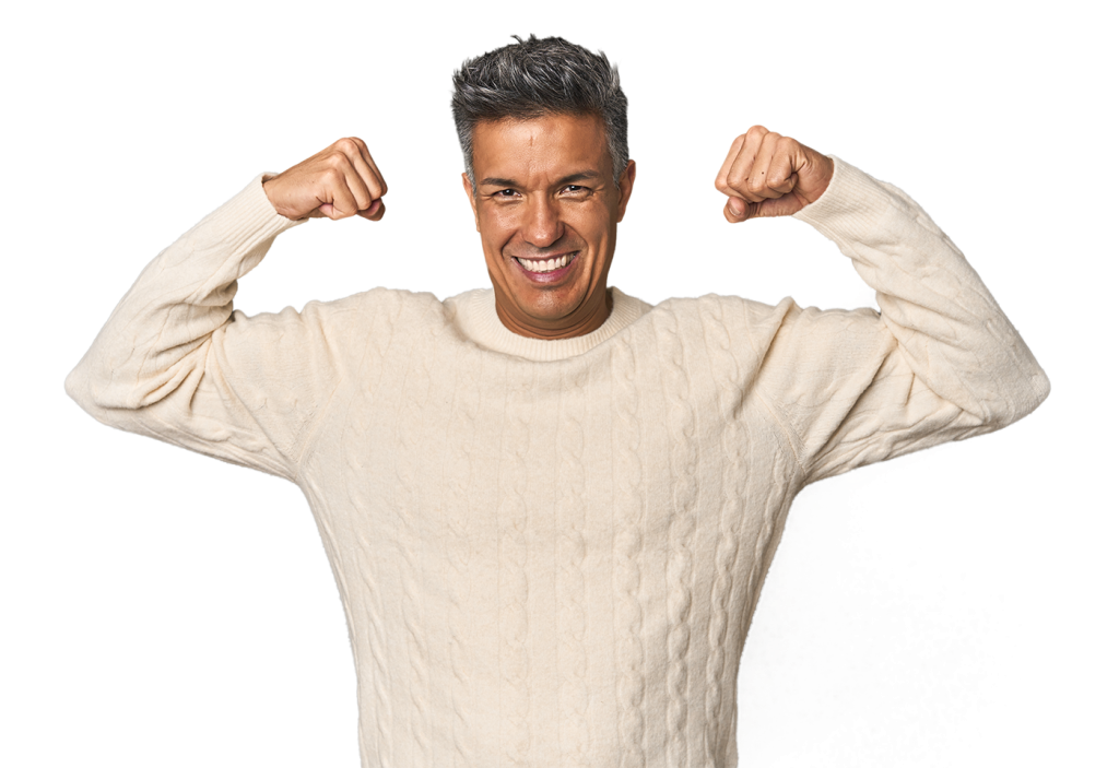 A man in a beige cable-knit sweater is smiling broadly and flexing his arms with both fists raised, displaying a confident and joyful demeanor. The background is white.